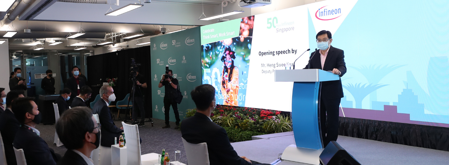 Man in suit with mask speaks at podium; Infineon logo, "Singapore" on screen behind him. Audience seated.